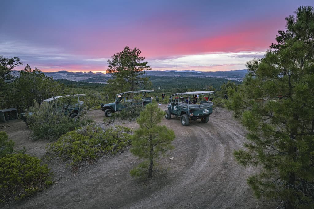 Off-road vehicles parked on a winding dirt path with a colorful sunset and mountains in the background.